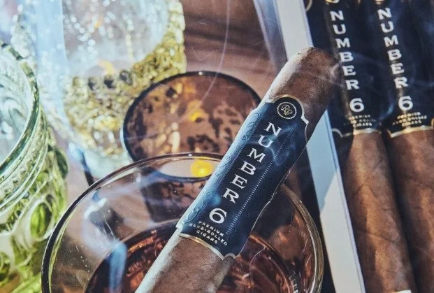 A cigar with a black band labeled 'NUMBER 6' resting on a glass of whiskey. In the background, there are other cigars and glass jars with decorative items.