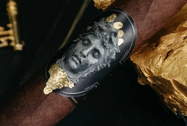 Close-up of a cigar band featuring a depiction of a classical Greek or Roman sculpture of a person's face with curly hair, decorated with gold accents.