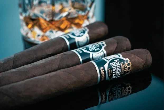 Three paired cigars with Kentucky Bastard labels resting on a reflective black surface, with a glass filled with ice and snacks in the background.