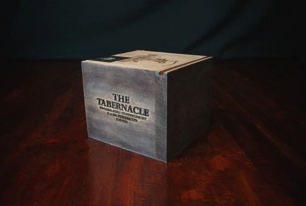 A cardboard box labeled 'The Tabernacle, The Assembly of the Called of God, Dothan, Alabama' sitting on a wooden surface.