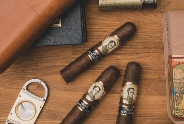 Four cigars with Fidel Castro's portrait and 'GRAN REPUBLICA' on bands, placed on a wooden surface among a gun holster, a lighter, and other objects.
