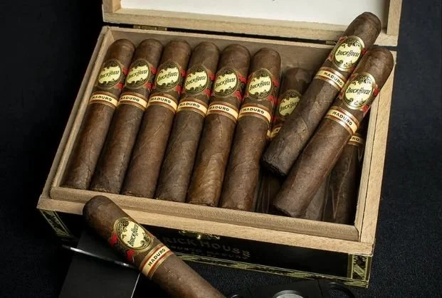 A wooden box filled with several cigars, some lying on top of others. The cigars have labels with the word "Maduro" and other branding details.