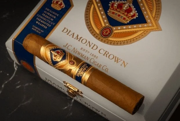 A Diamond Crown cigar with a brown wrapper resting on a box of Diamond Crown cigars. The box features a crown emblem and gold and blue design elements.