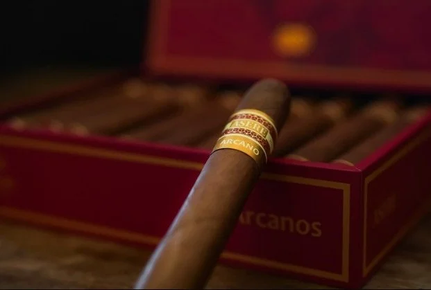 A cigar resting on the edge of a red box of cigars on a wooden surface.