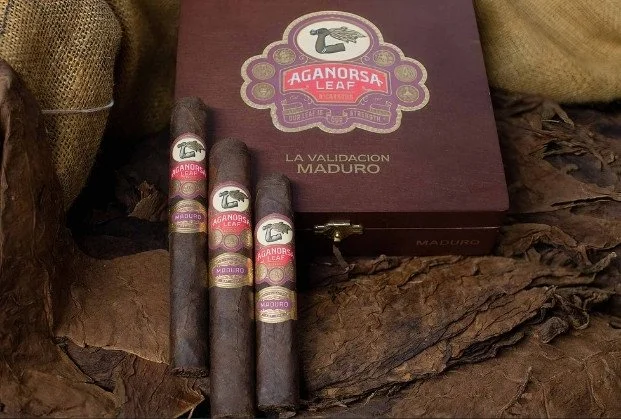 Three cigars with bands reading "Aganorsa Leaf Maduro", placed in front of a wooden box with similar branding, all set on a rustic wooden surface.