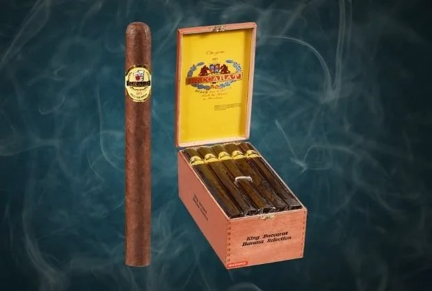 A cigar standing next to an open box of cigars with a yellow interior and a pink exterior labeled 'King Cameroon Havana Selection'.