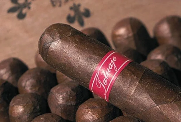 A close-up view of a cigar labeled 'Tajagne' resting on a wooden surface surrounded by additional cigars.