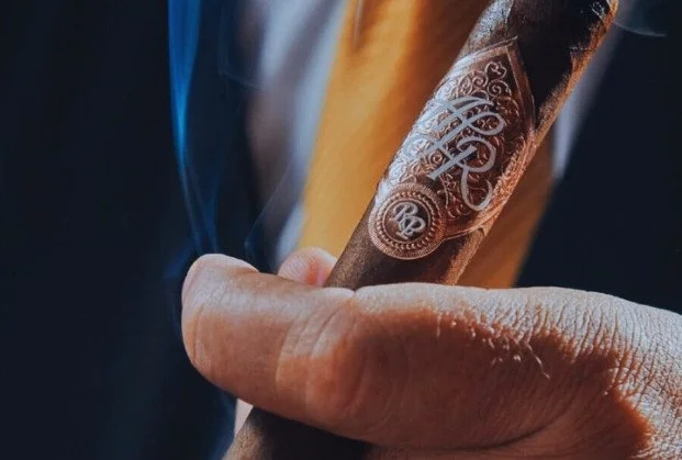 Close-up of a hand holding a lit cigar with decorative branding, smoke rising from the cigar.