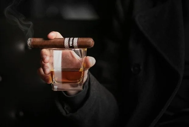 Person holding a glass of whiskey with a lit cigar resting on the rim.
