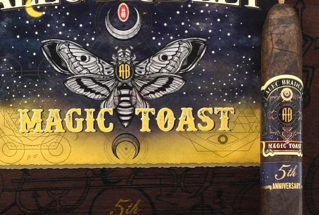 A cigar with a band that reads "Alec Bradley Magic Toast 5th Anniversary" positioned next to a box with a matching design and text.
