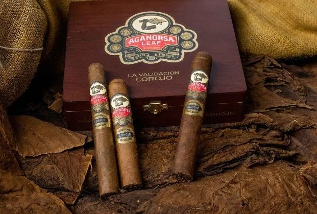 Four cigars with labels placed in front of a wooden box labeled "Aganorsa Leaf" and "La Validacion Corojo". The cigars are surrounded by dried tobacco leaves.