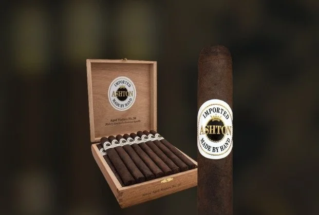 Open wooden box of Ashton cigars and a single dark brown cigar with a white and gold Ashton label.