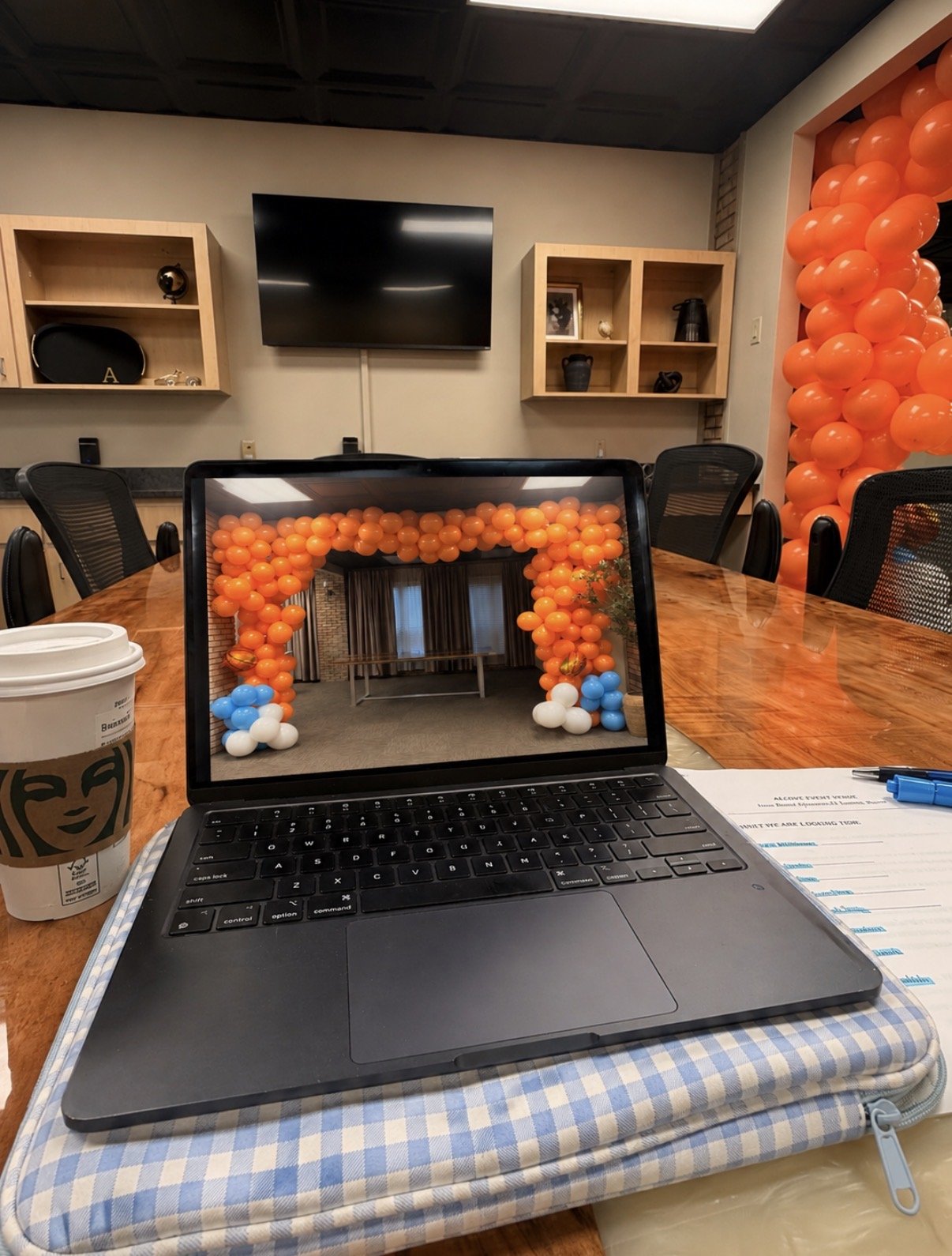 Conference room decorated with orange balloons creating an arch and large column. A table with chairs is in the room. The laptop screen shows the same decorated space as a reflection. Items on the table include a Starbucks coffee cup and documents.