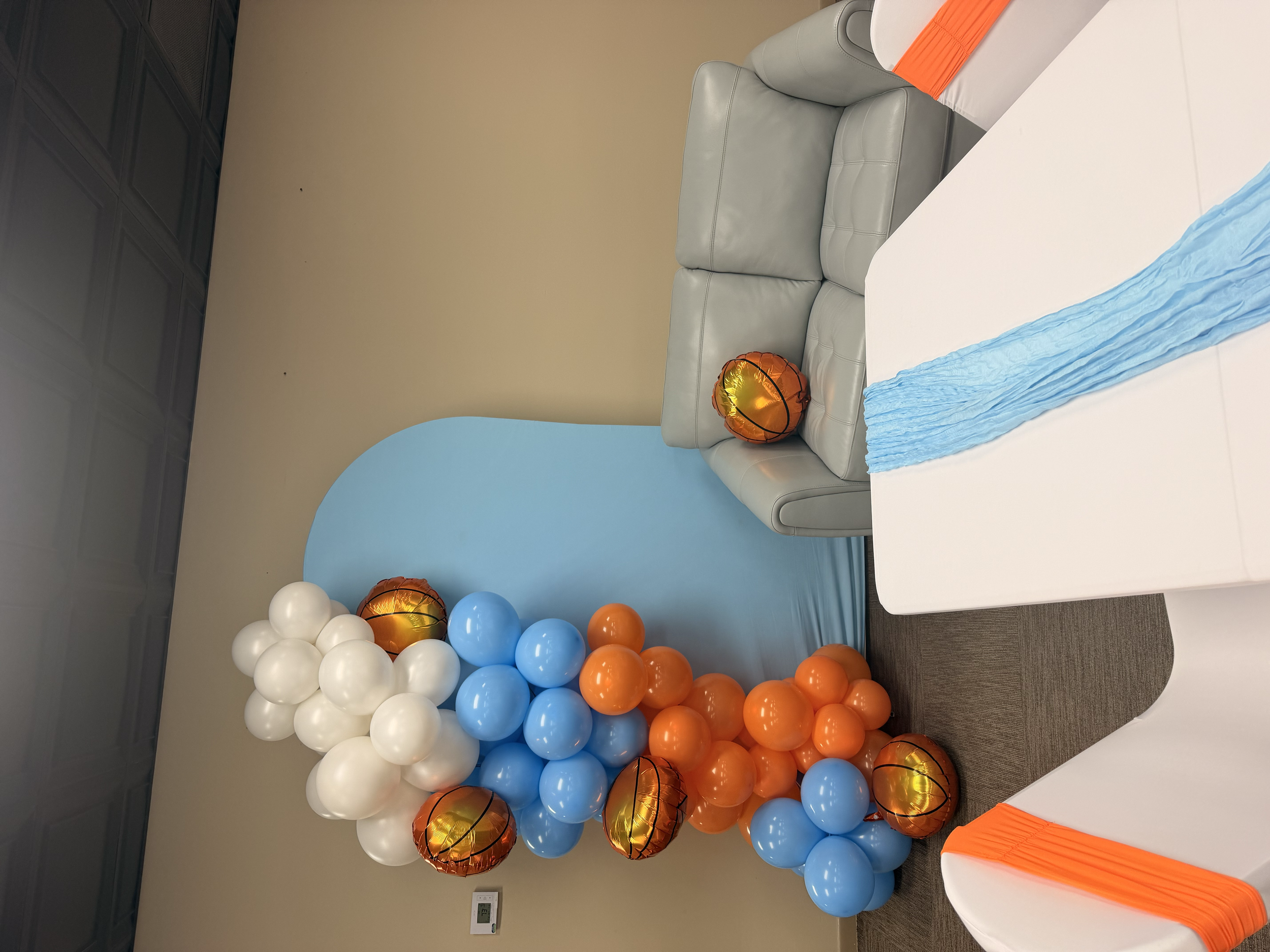 Balloon decorations with white, blue, and orange balloons arranged in clusters, set up for a celebration or party in a room with beige walls, a blue accent wall, and a table decorated with an orange overlay.