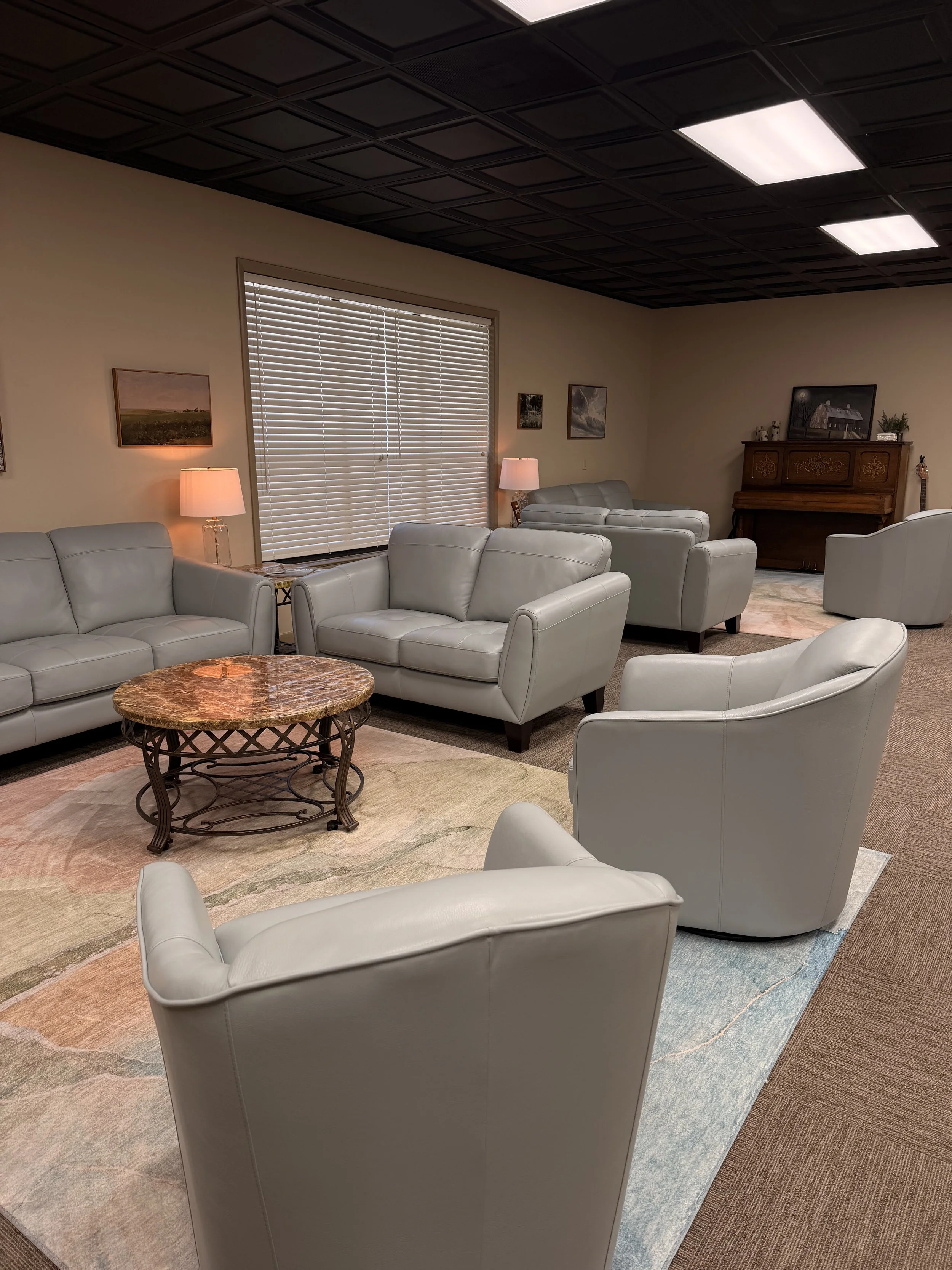 A living room with beige walls, white leather sofa set, armchairs, a wooden coffee table, a rug, window with blinds, lamps, and a piano, decorated with various pictures and small decorative items.