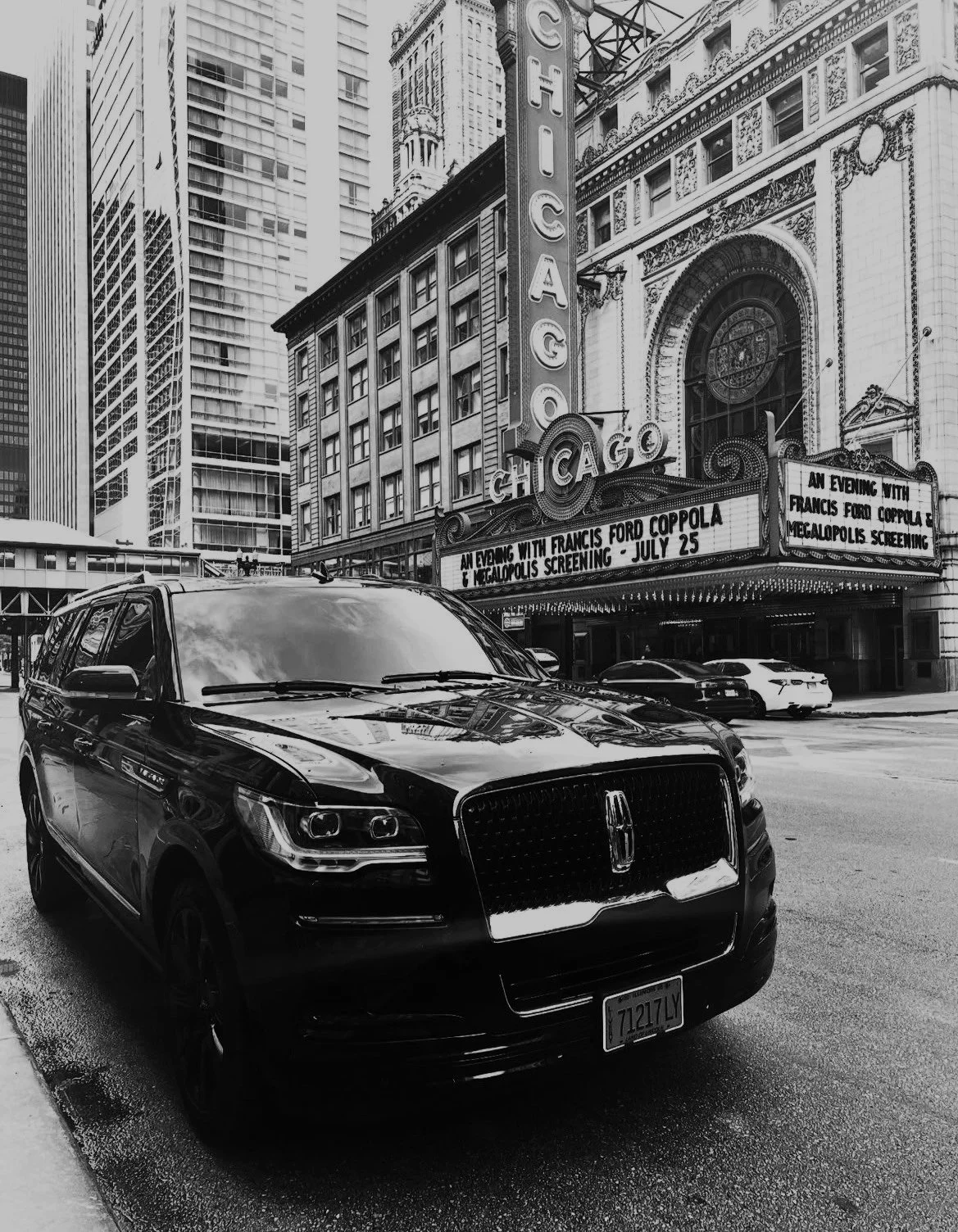 Black SUV parked in front of the Chicago Theatre marquee and sign in downtown Chicago, with tall buildings behind.