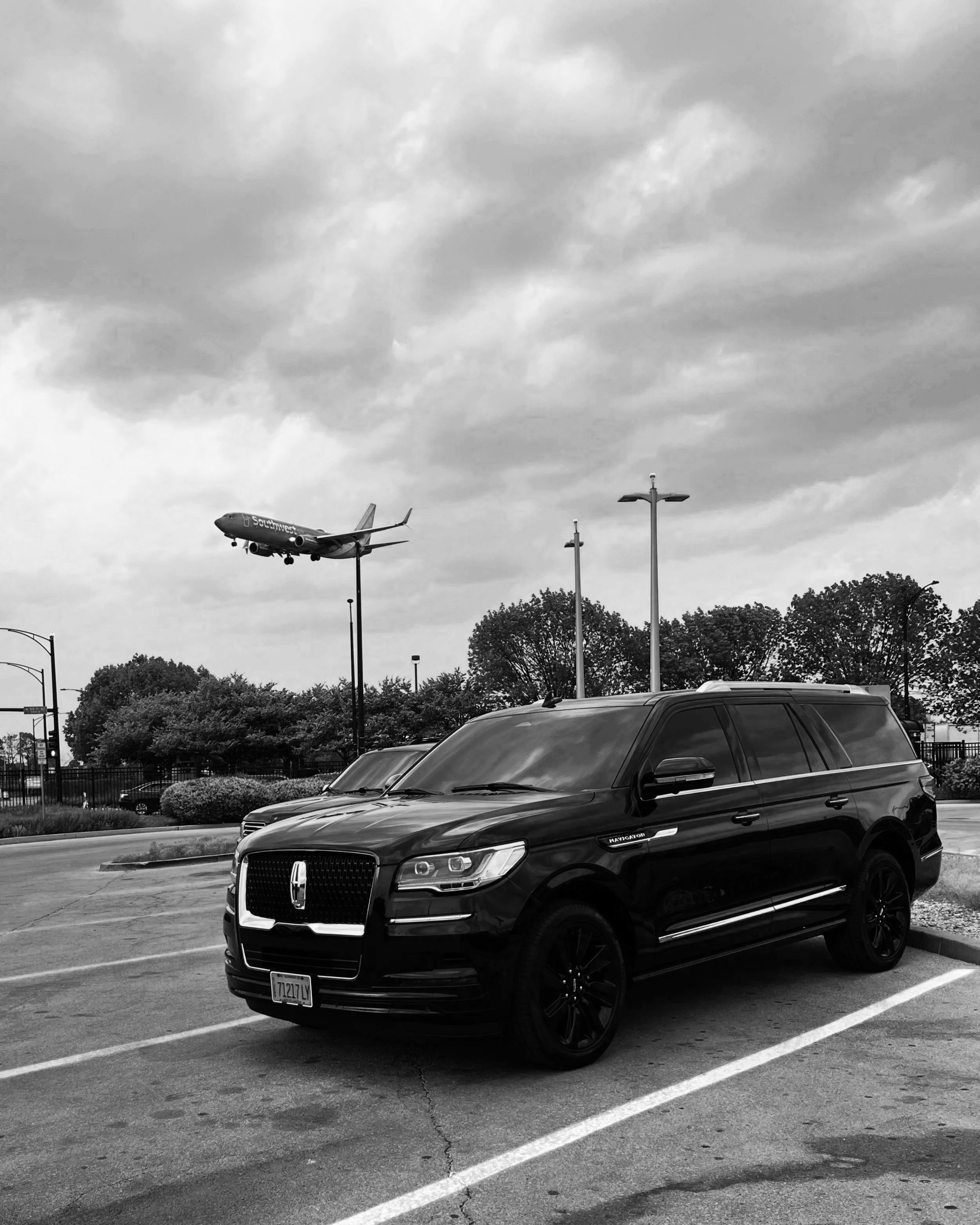 Black Lincoln Navigator SUV parked in an outdoor lot with trees and streetlights, while a Southwest Airlines airplane is flying in the cloudy sky overhead.