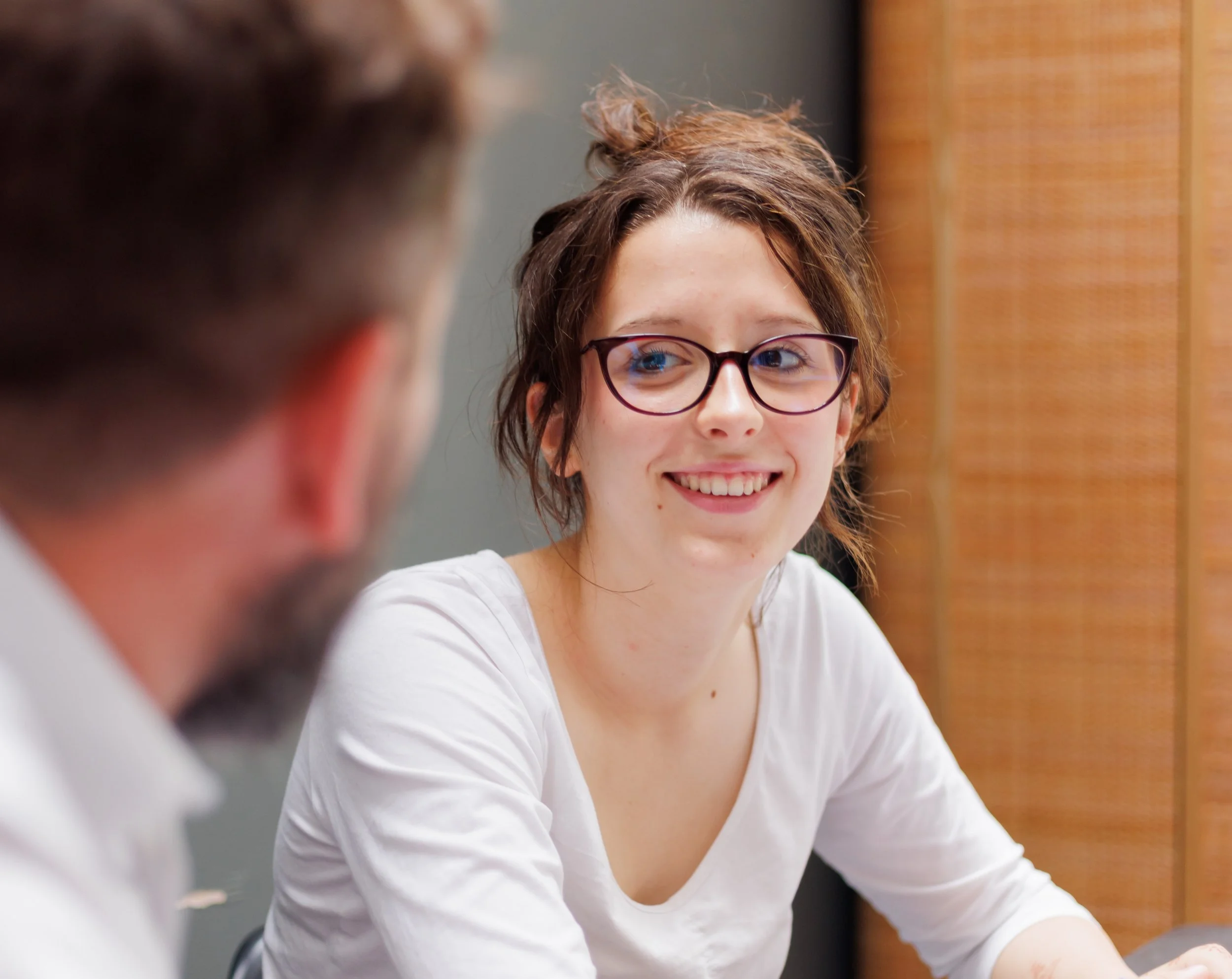 A young woman with glasses and wavy brown hair smiling while talking to a man with a beard, seen from behind, in an indoor setting with a wooden wall.