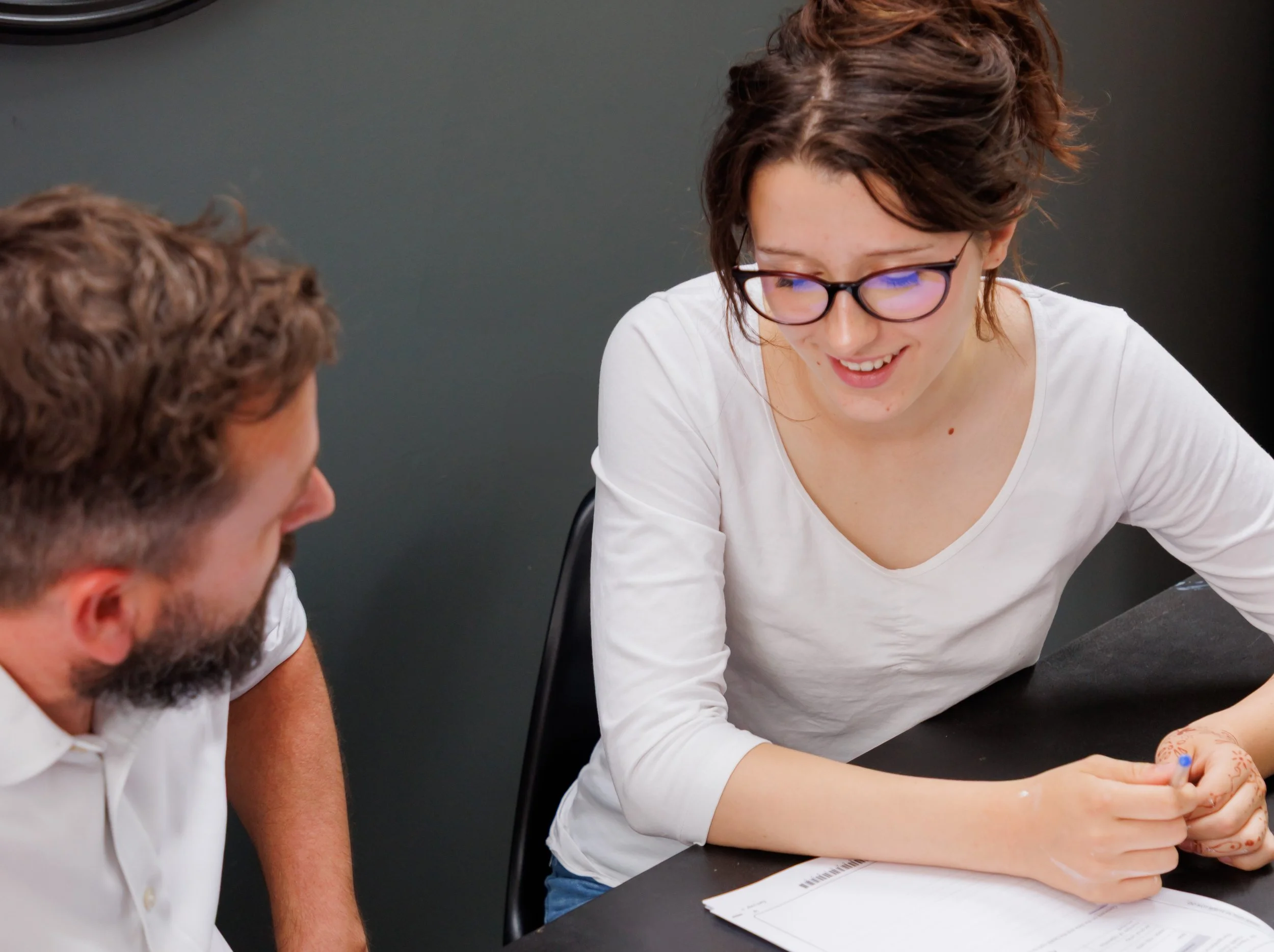 A young woman with glasses and short brown hair smiling and talking to a bearded man with curly hair in an office setting, sitting at a table with papers and a pen.