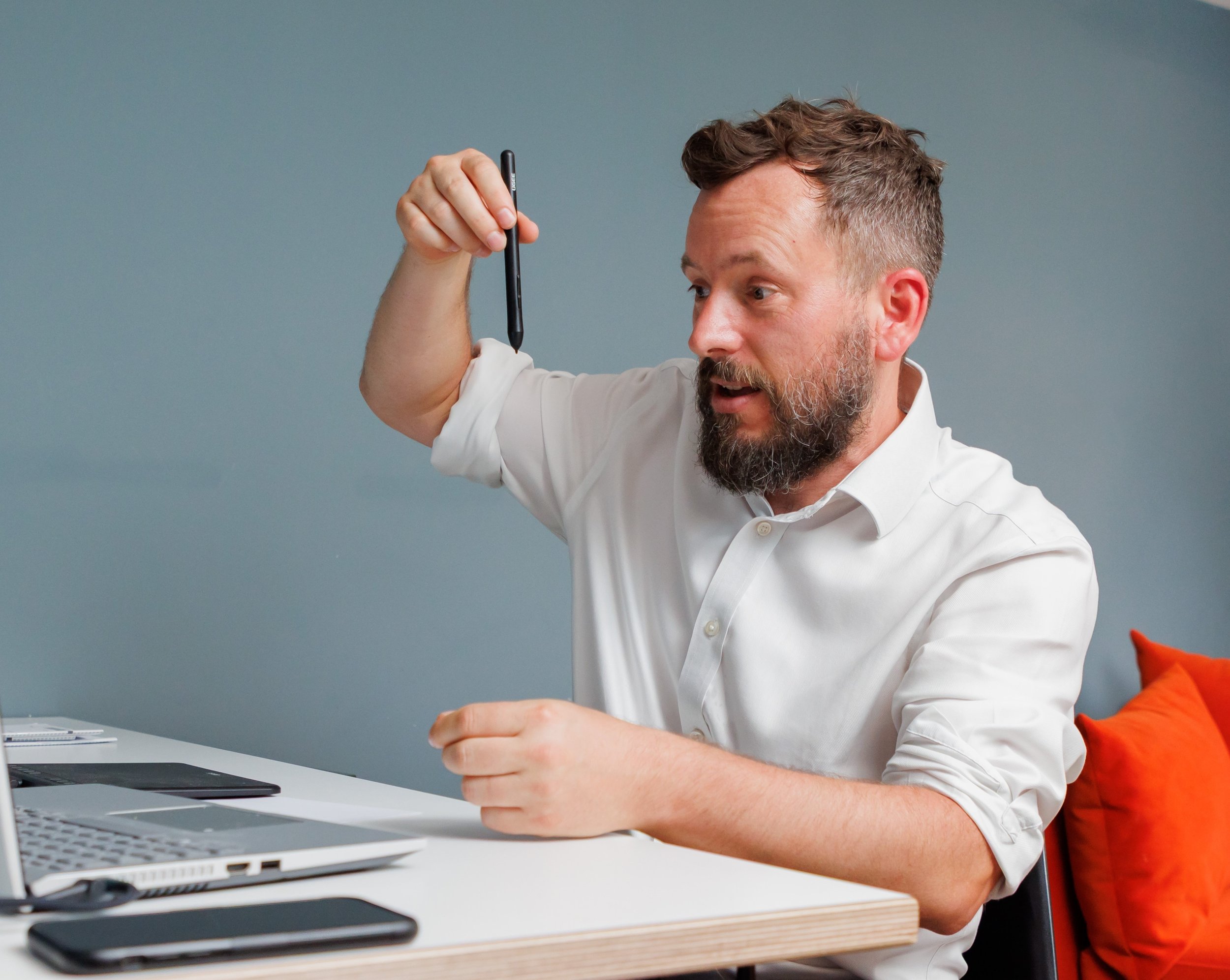 Man with a beard and short hair in a white shirt holding a stylus or pen, appearing to perform a demonstration or presentation, sitting at a desk with electronic devices, in front of a blue-gray wall.