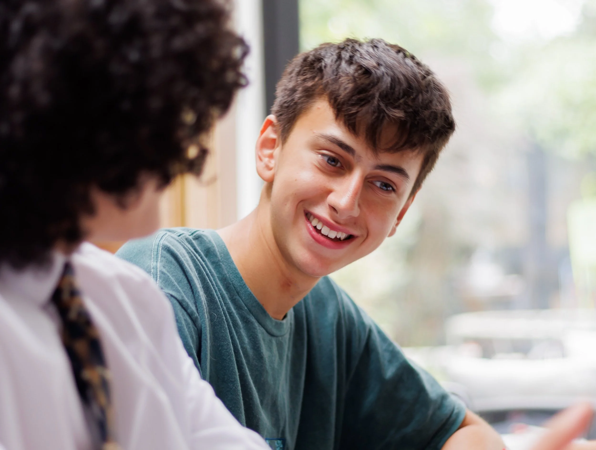 A young man and a woman with curly hair having a conversation indoors near a window.