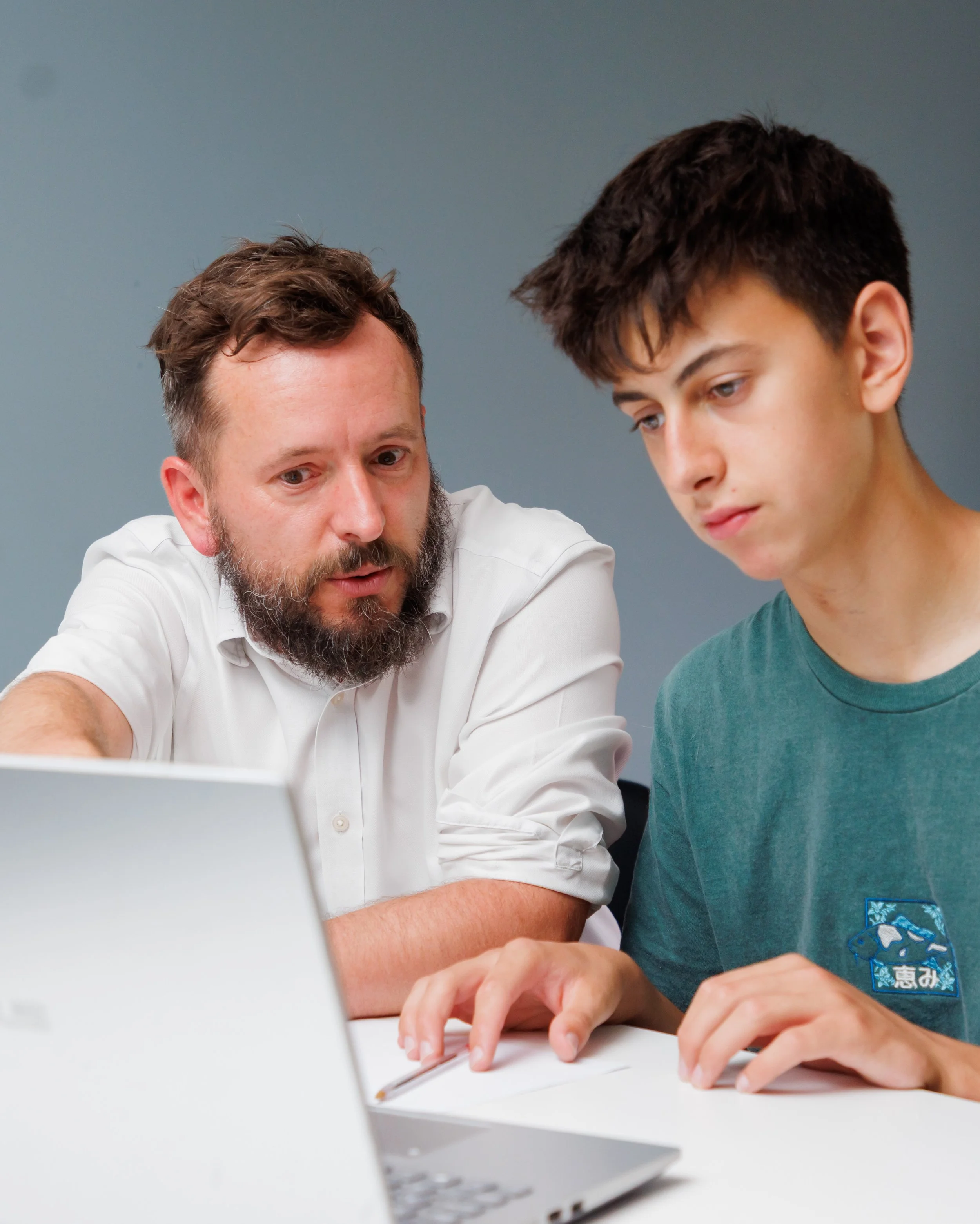 Man and teenager using a laptop together in a room with a plain gray background.
