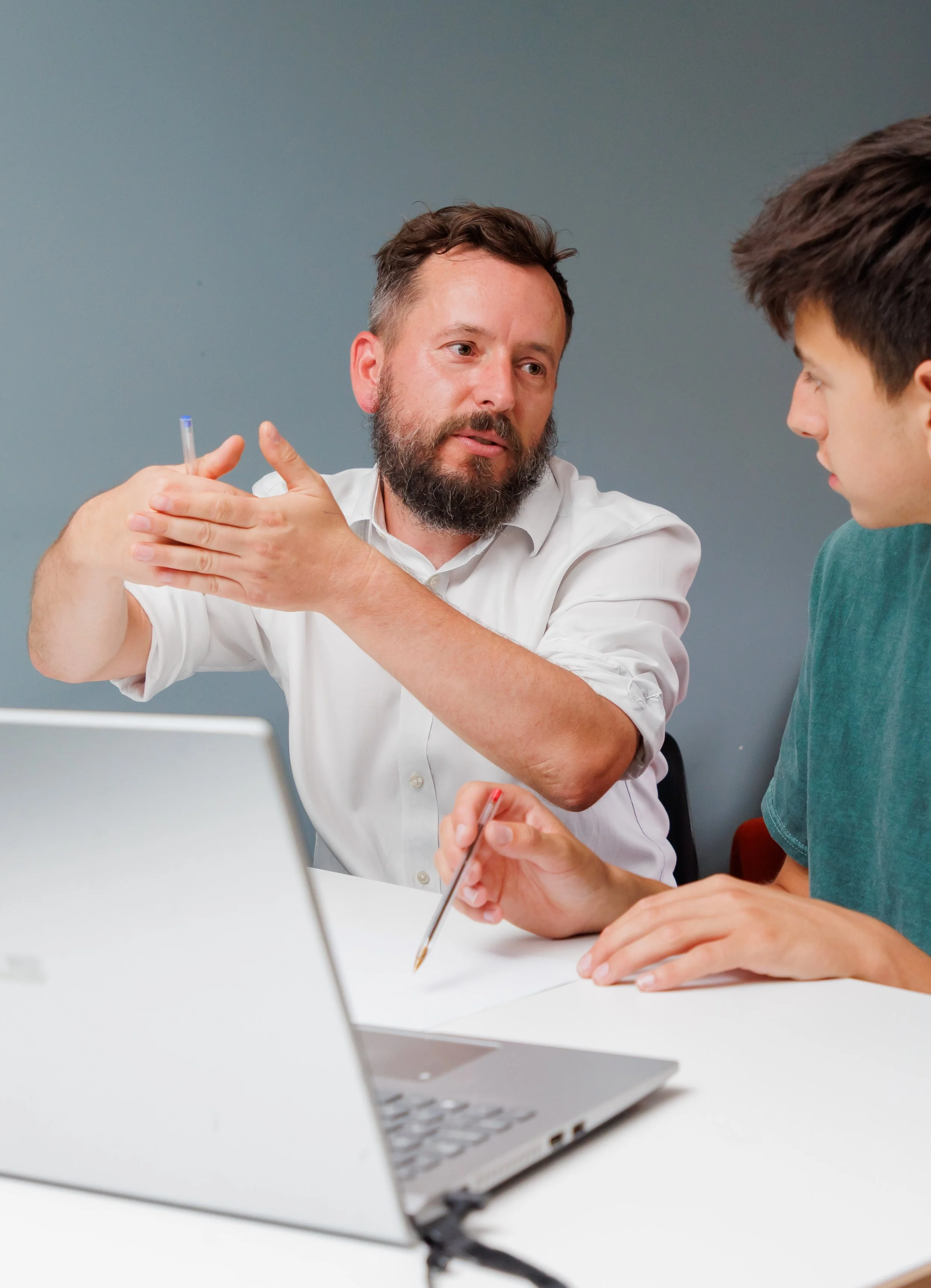 A man with a beard in a white shirt explaining something to a young man in a teal shirt at a desk with a laptop and papers.