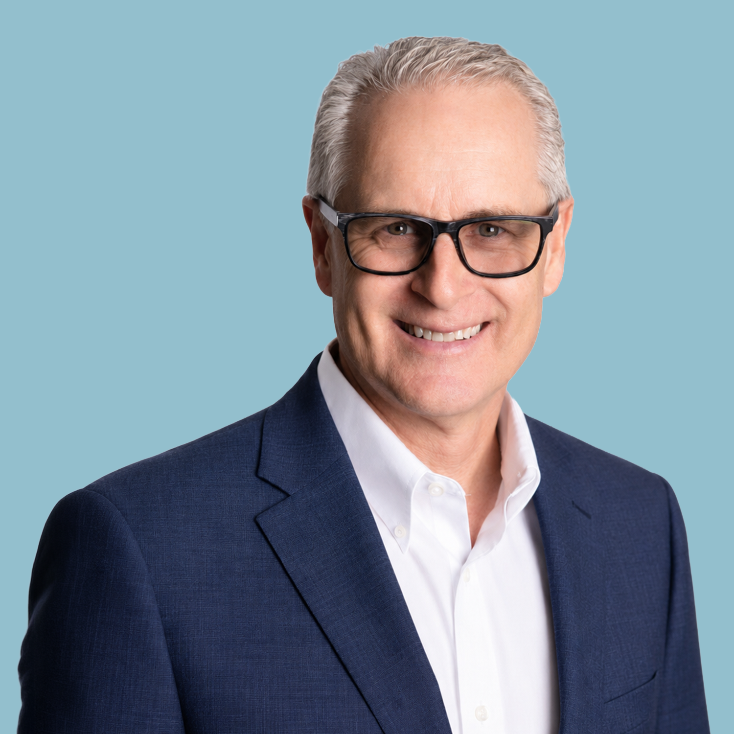Portrait of a middle-aged man with short, light-colored hair, wearing a light blue patterned dress shirt, standing against a plain white background.