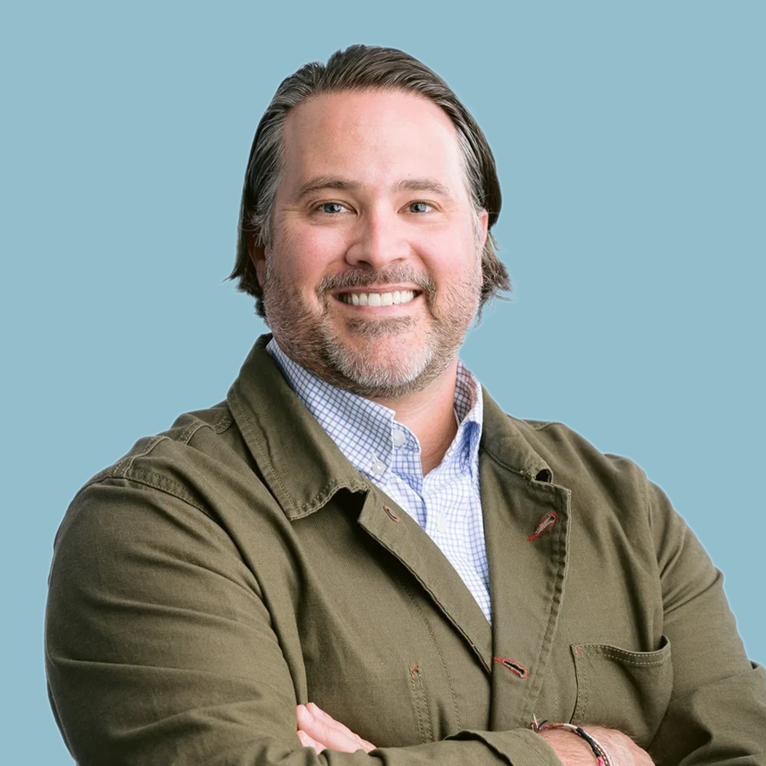 A smiling man with gray hair and a beard, wearing a khaki jacket over a blue checkered shirt, standing against a plain white background.
