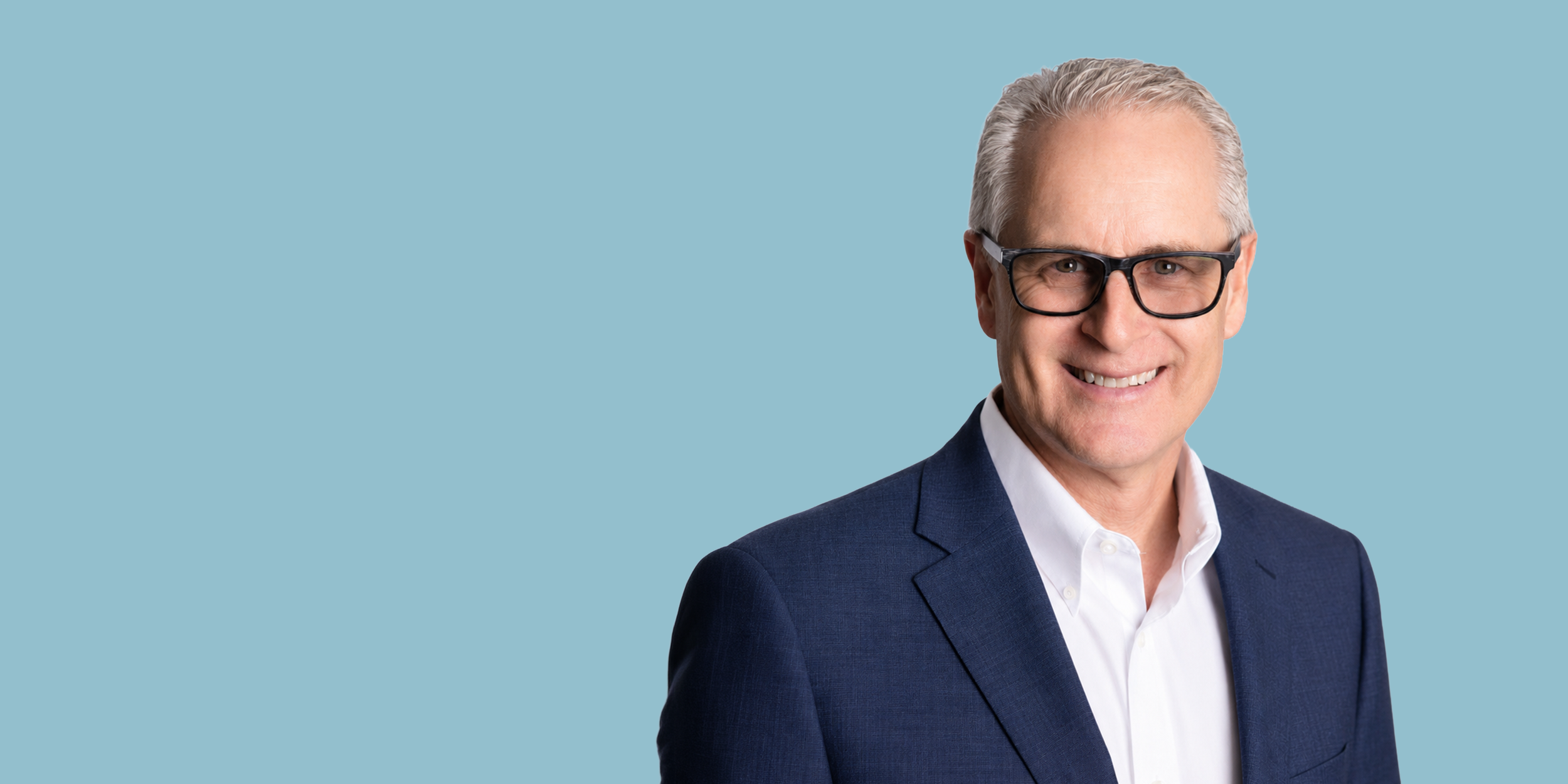 Portrait of a middle-aged man with gray hair, glasses, wearing a navy blue suit and white shirt, smiling against a light blue background.