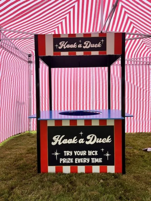 Red and white striped tent with a game booth labeled "Hook a Duck" offering a prize for every attempt, featuring a blue basin with floating ducks.