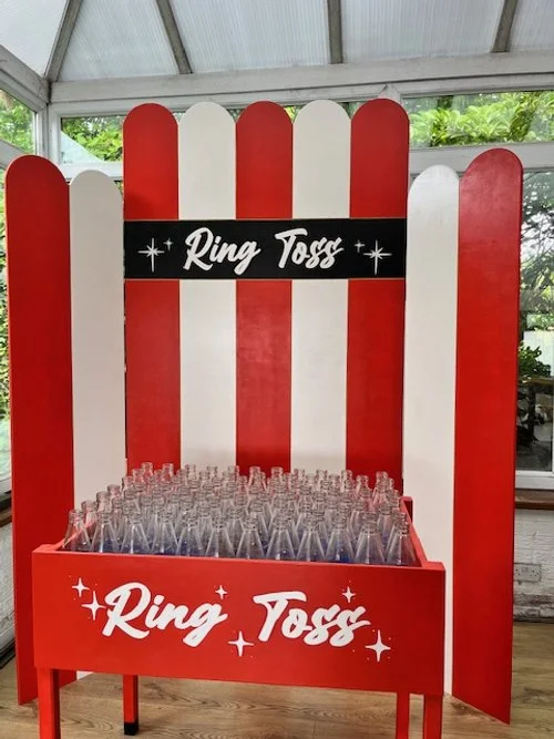 A red and white striped wooden fence with a sign reading "Ring Toss" in the middle, and a table in front filled with glass bottles for a ring toss game.