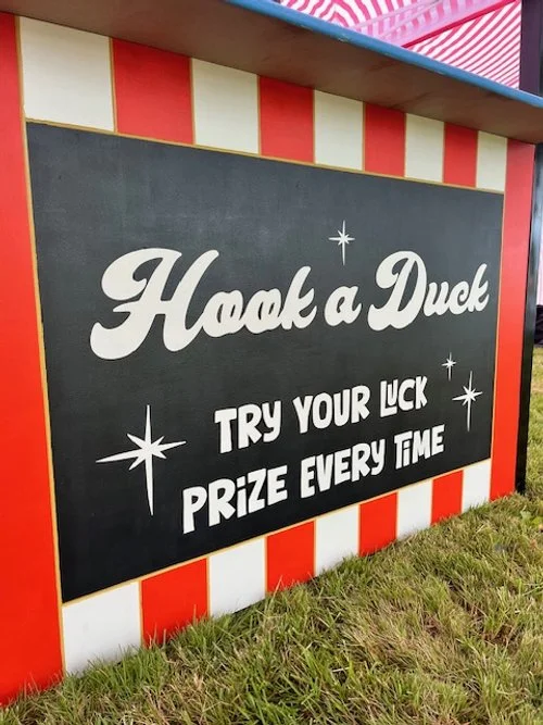 A game booth sign reading 'Hook a Duck' with instructions to try your luck and prize every time, decorated with red and white stripes.