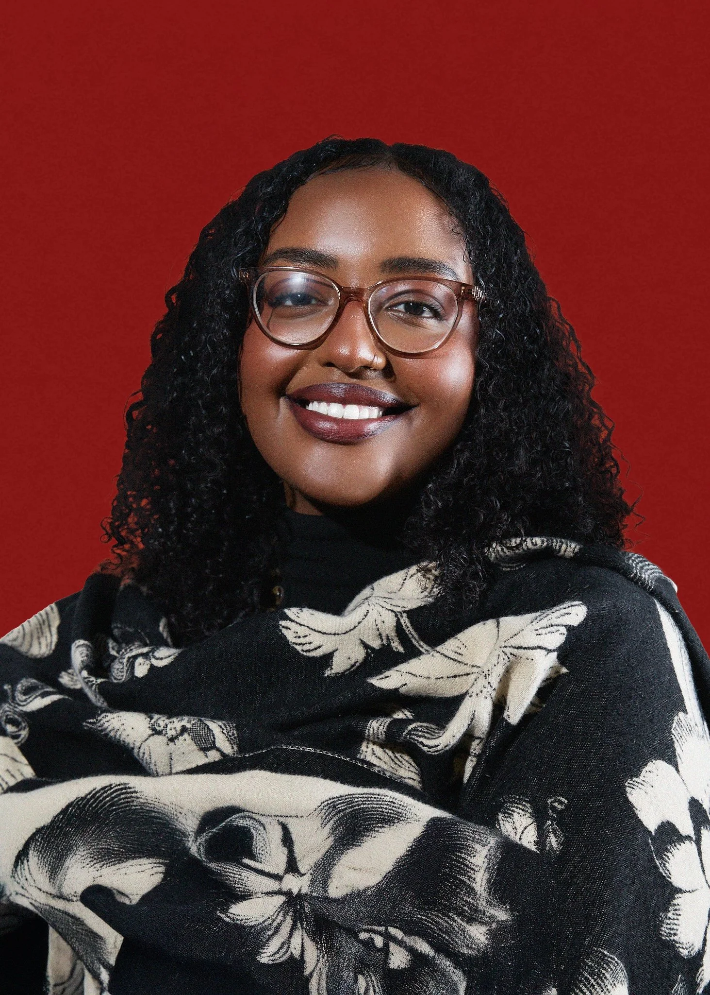 A smiling woman with curly black hair, wearing glasses and a black sweater with a cream-colored floral shawl, standing in front of a red background.