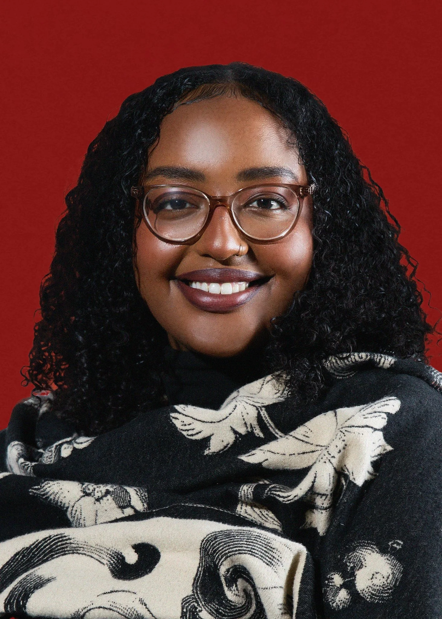 Portrait of a smiling Black woman with curly hair, wearing glasses, and a black and white patterned scarf against a red background.