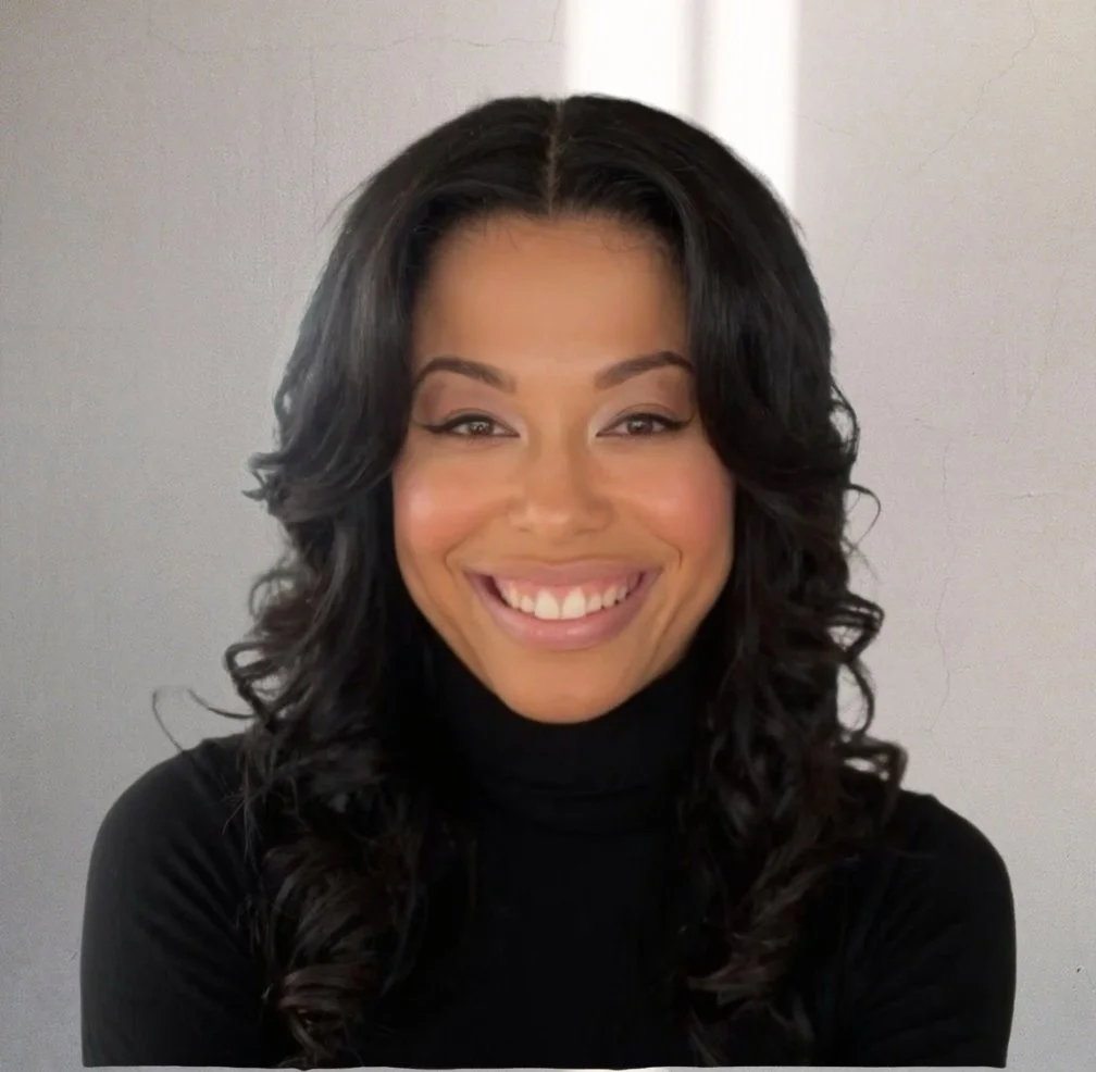A young woman with long dark hair, smiling, wearing a yellow cardigan over a white top, seated against a plain wall.