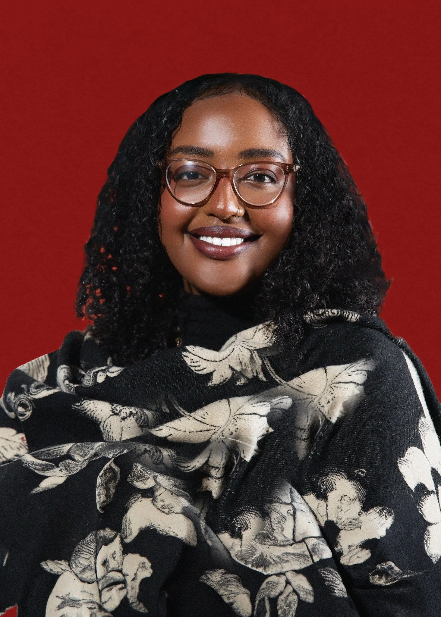 Portrait of a woman with glasses and curly hair, smiling, wearing a black and white floral scarf against a red background.