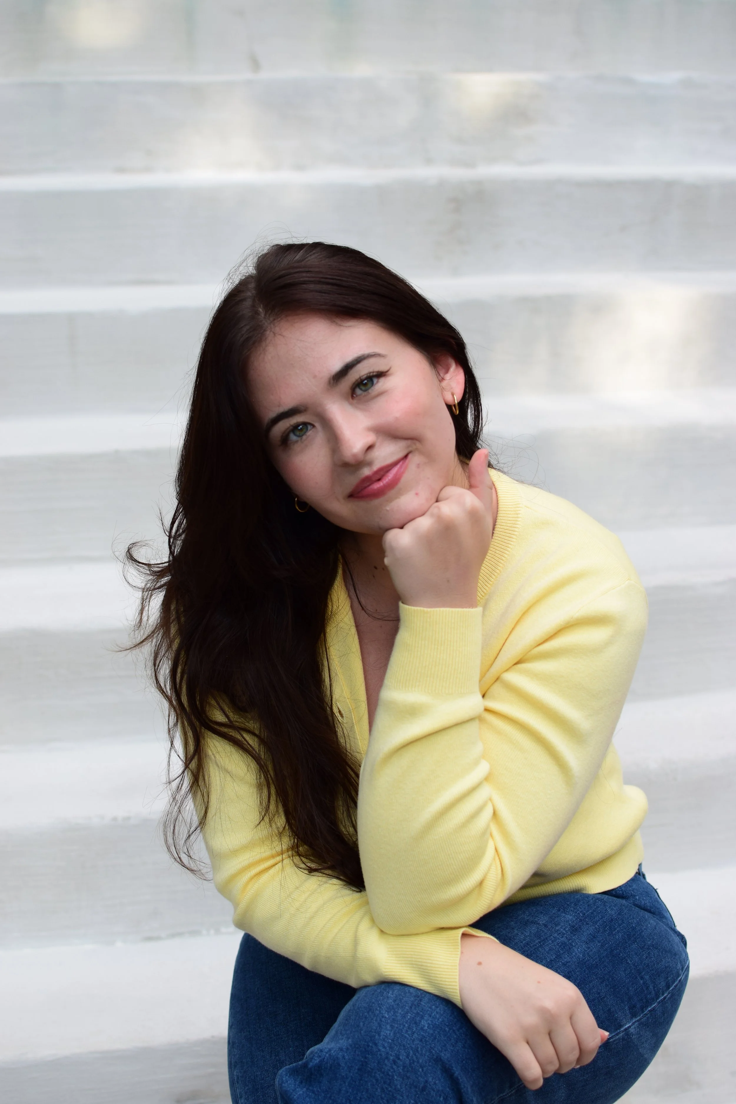 A woman with long dark hair, wearing a yellow sweater and blue jeans, sitting on white stairs.