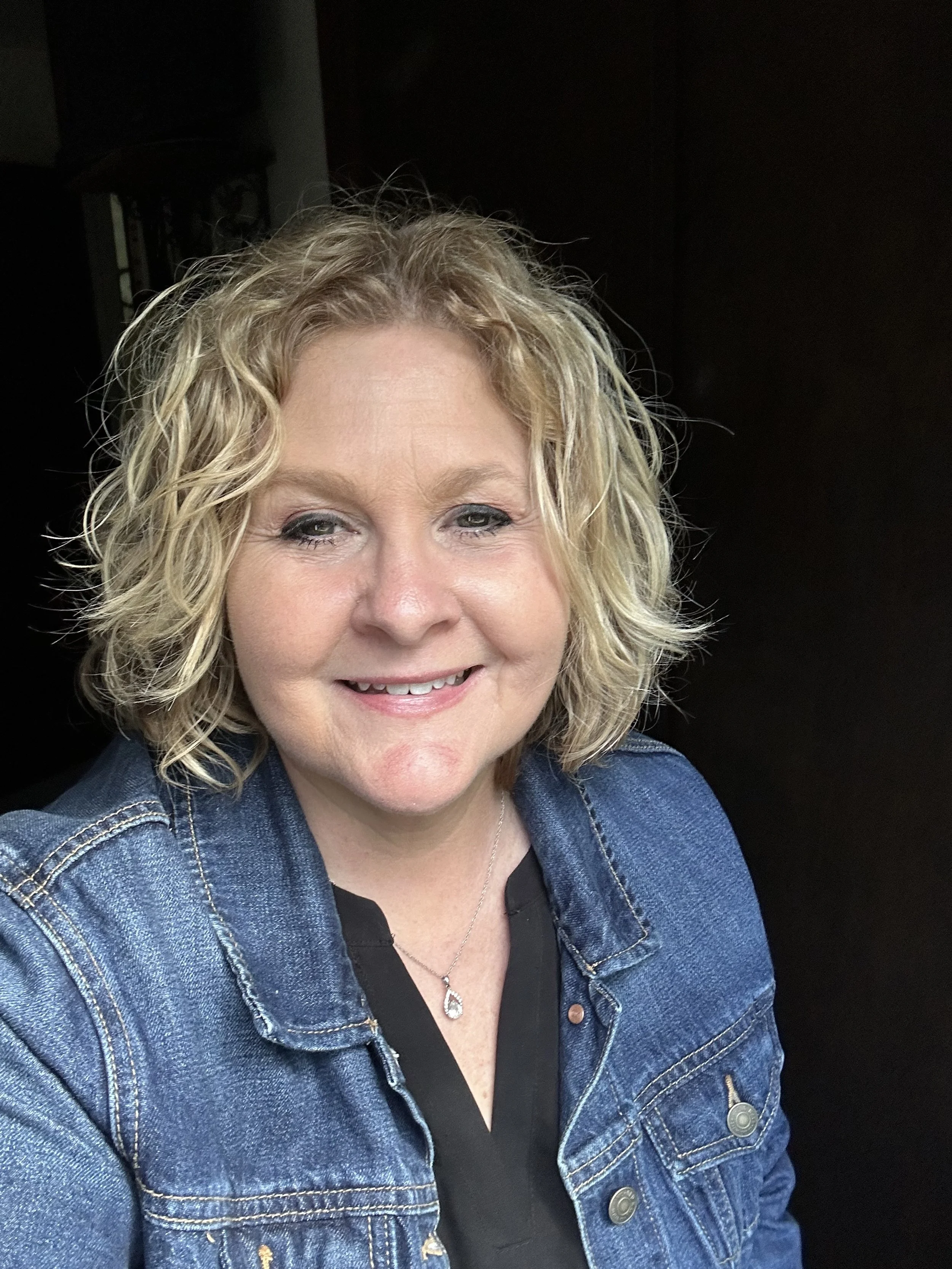 A woman with curly blonde hair smiling, wearing a denim jacket and a black top, with a silver necklace.