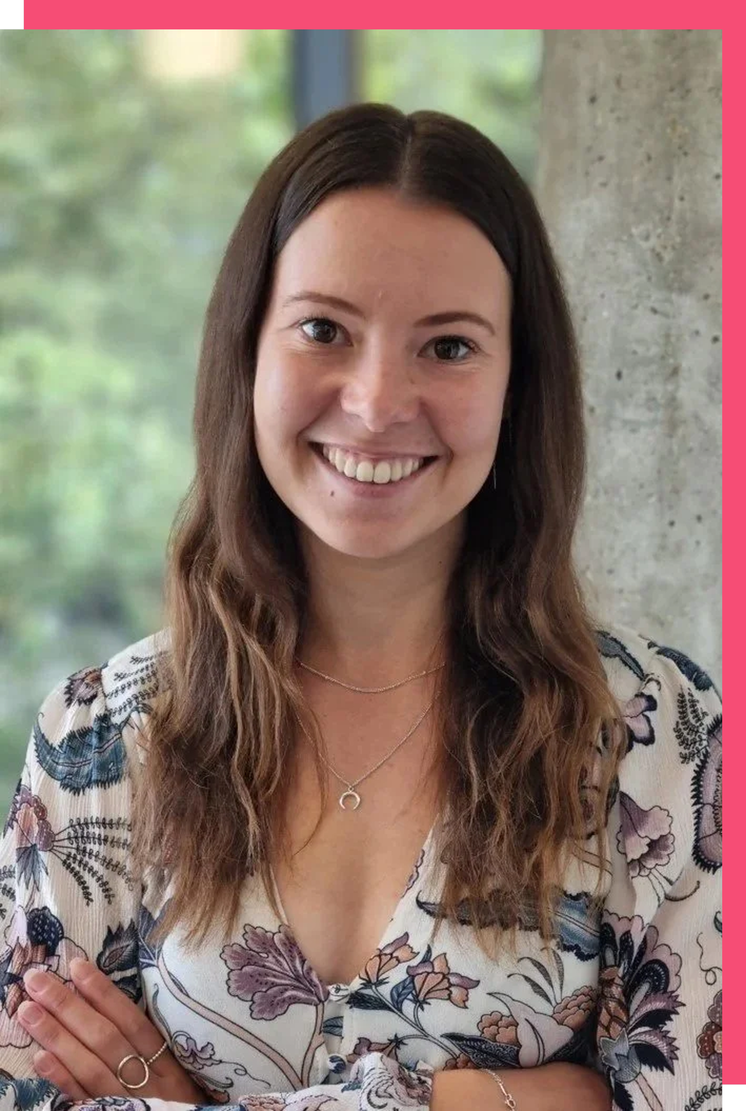 A woman with long brown hair, smiling, wearing a patterned blouse with floral designs, standing indoors near a concrete wall with a window showing greenery outside.