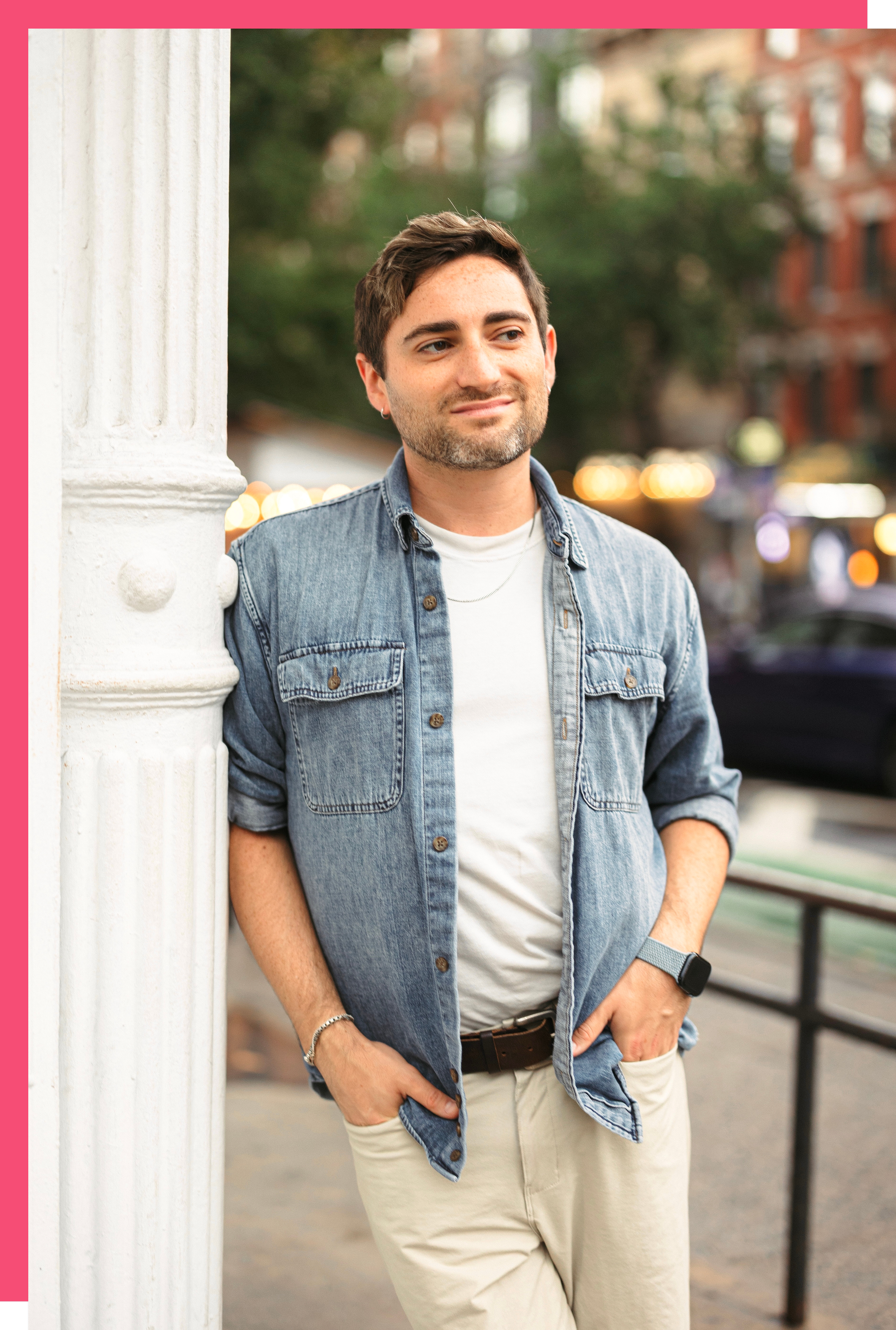 A young man with dark hair and a beard standing outdoors against a white architectural column, wearing a denim jacket, white shirt, beige pants, and a smartwatch, with blurred background of cityscape and lights.