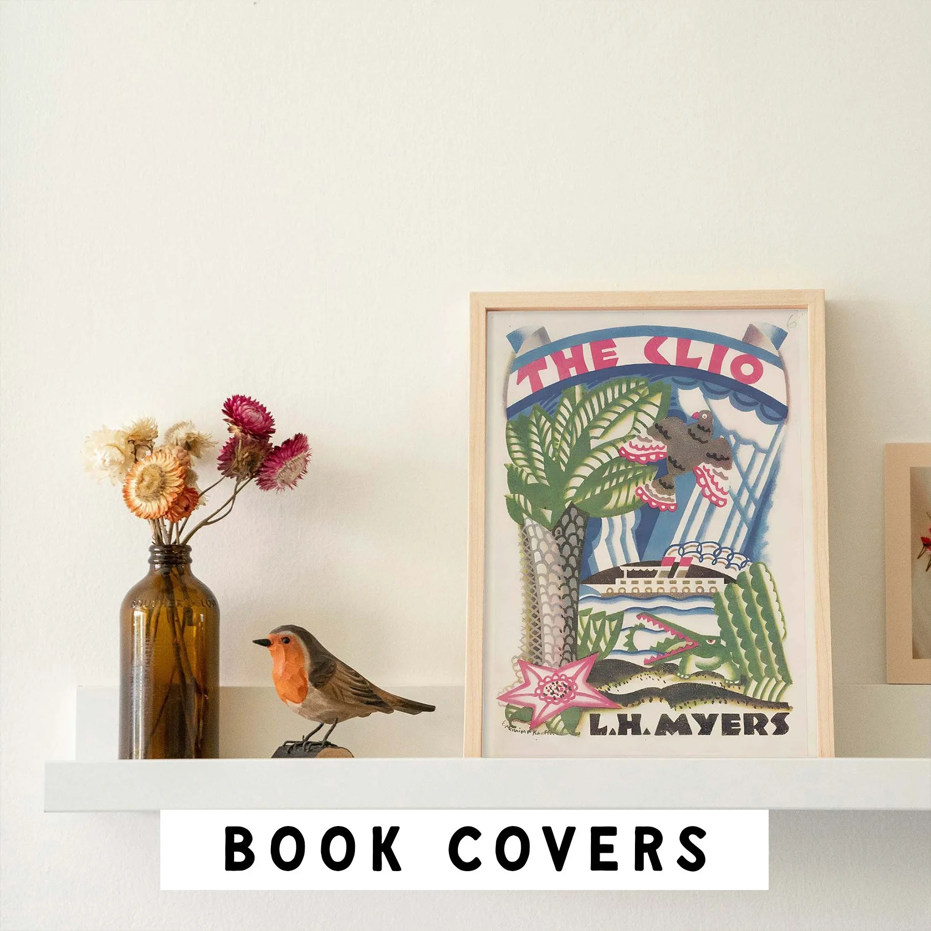 Decorative wall display featuring a vase with dried flowers, a bird figurine, a framed illustrated poster, and a sign reading 'BOOK COVERS'.