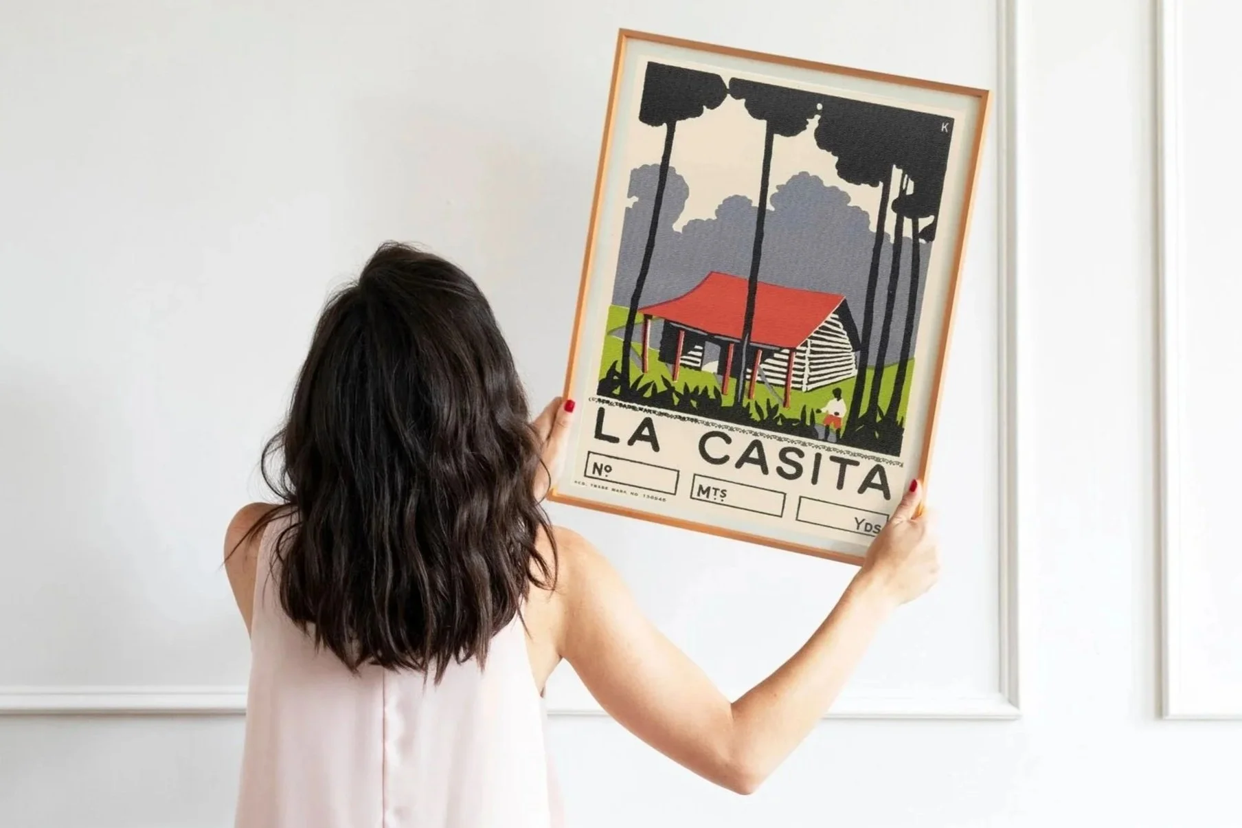 Woman with long dark hair holding a framed illustration of a house on stilts with a red roof, black stilts, and green grass, with a background of dark clouds and trees.