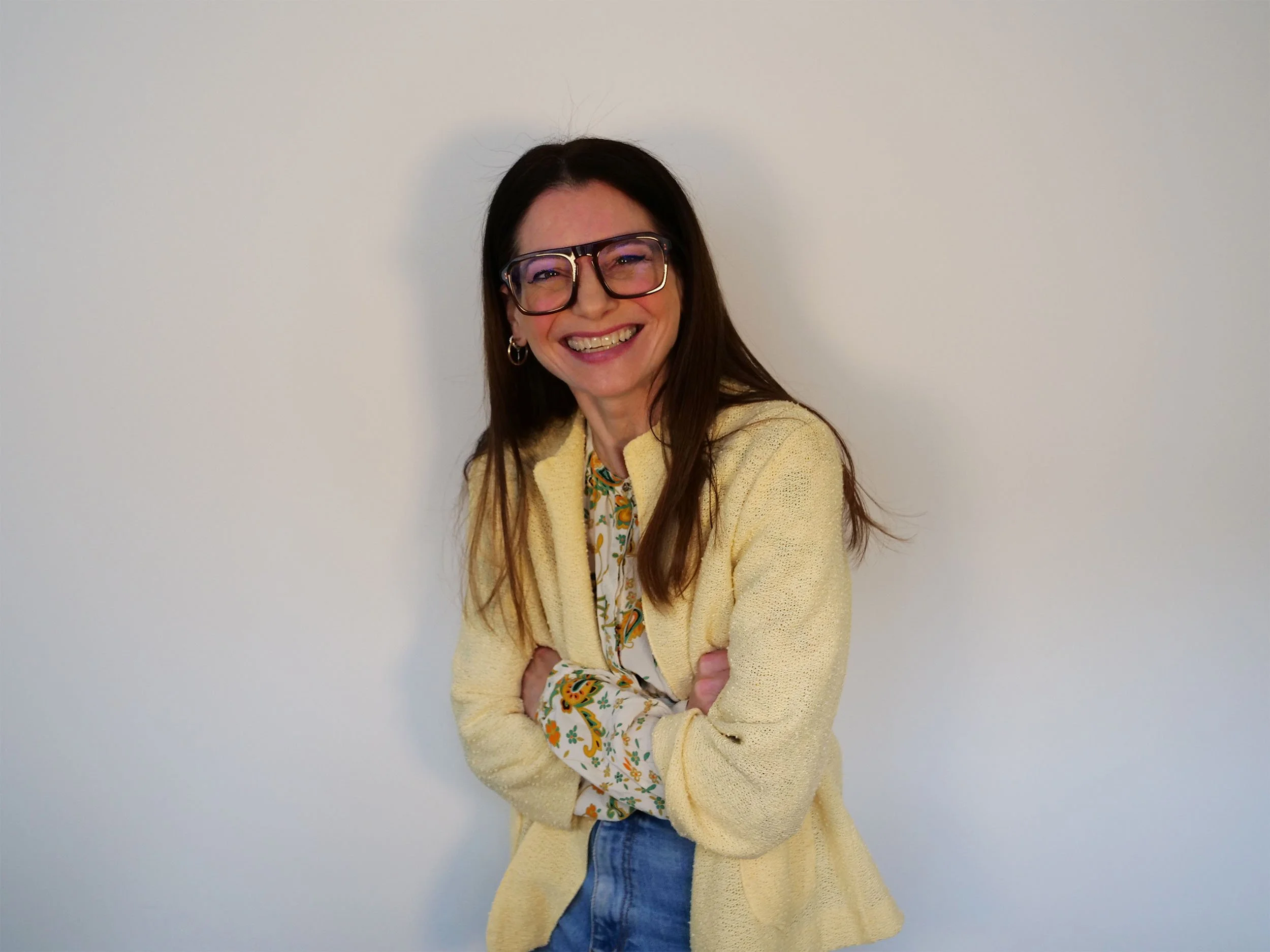 A woman with long brown hair, glasses, and hoop earrings is smiling and posing with her arms crossed in front of a plain white wall. She is wearing a yellow vintage cardigan over a floral patterned shirt and blue jeans.
