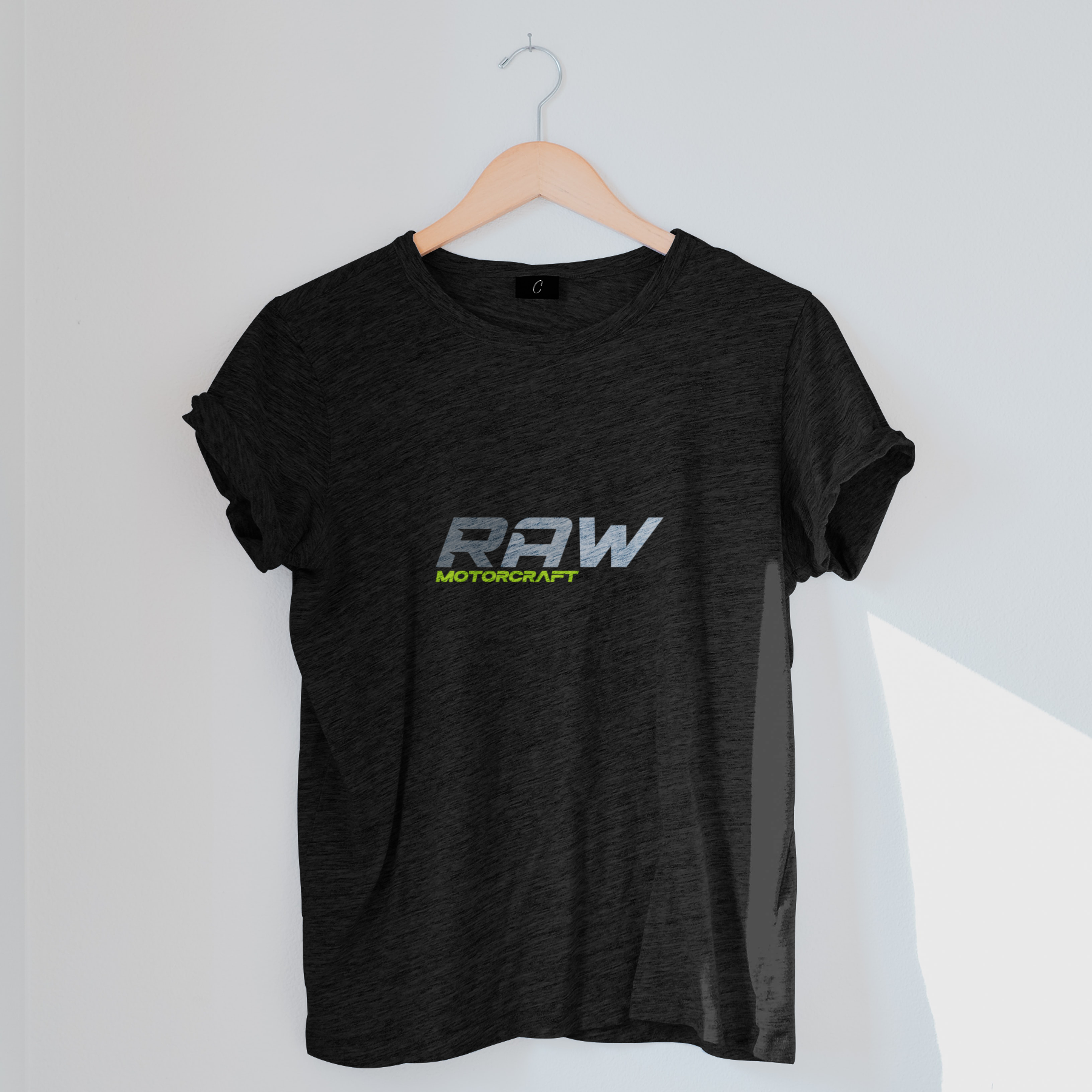 Black T-shirt hanging on a wooden hanger against a white wall, featuring a logo with the words "RAW MOTORCRAFT" in stylized font.