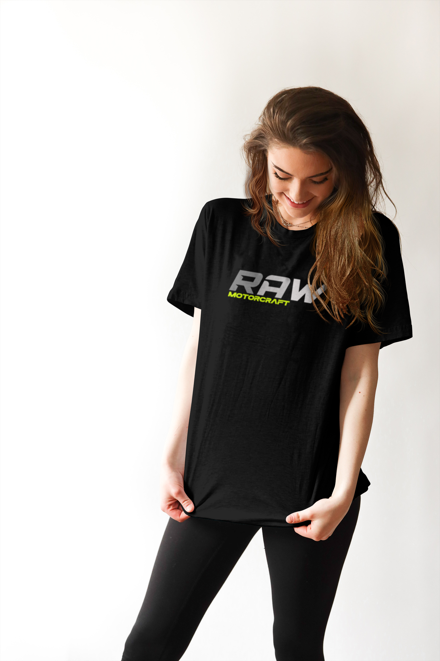 A young woman with long brown hair wearing a black t-shirt with "RAW MOTORCRAFT" printed on it, smiling and looking down, standing against a plain white background.