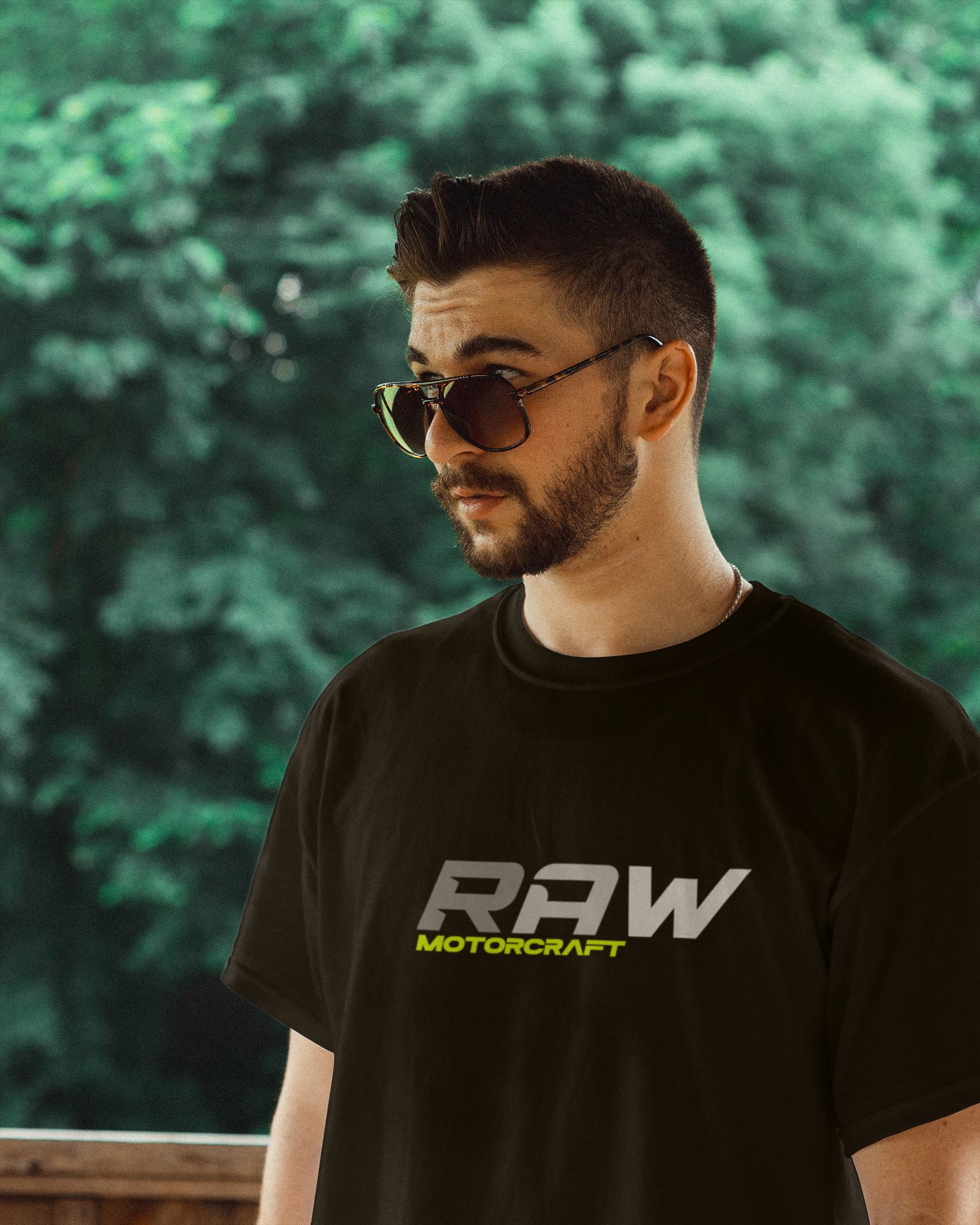 A man wearing sunglasses and a black T-shirt with the words 'RAW MOTORCRAFT' printed on it, standing outdoors with a blurred green forest background.