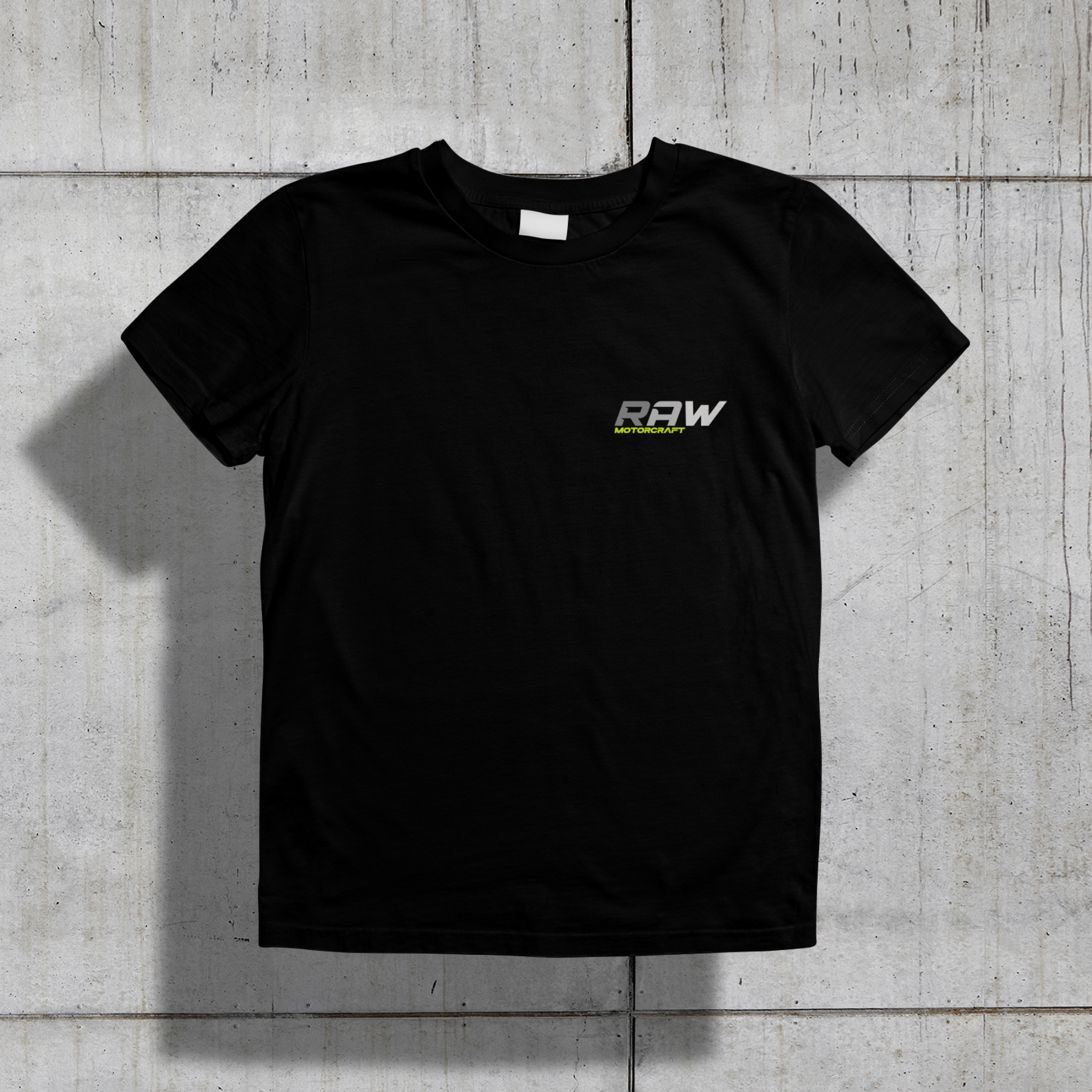 Black T-shirt with 'RAW MOTORCRAFT' logo in gray and lime green on the front, laid flat on a concrete surface.