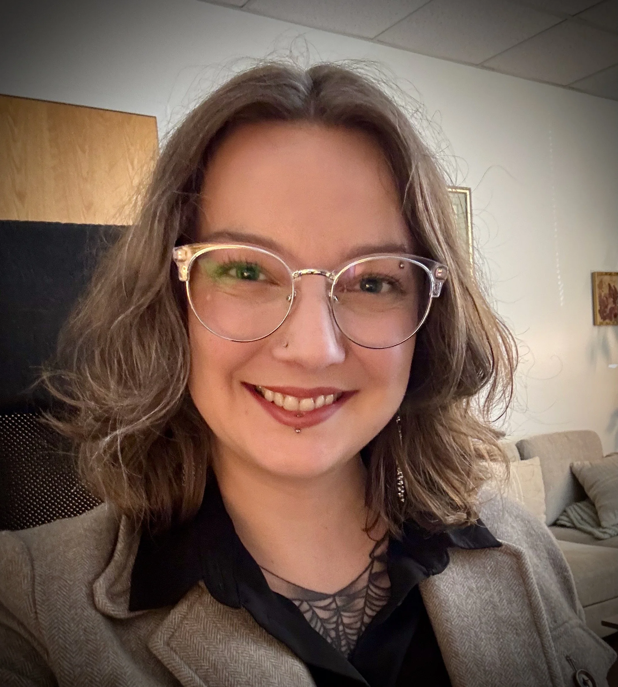 A smiling woman with shoulder-length wavy brown hair wearing glasses, a nose piercing, and a labret piercing. She is dressed in a blazer and shirt, and is seated inside a room with beige couches and framed pictures on the wall.