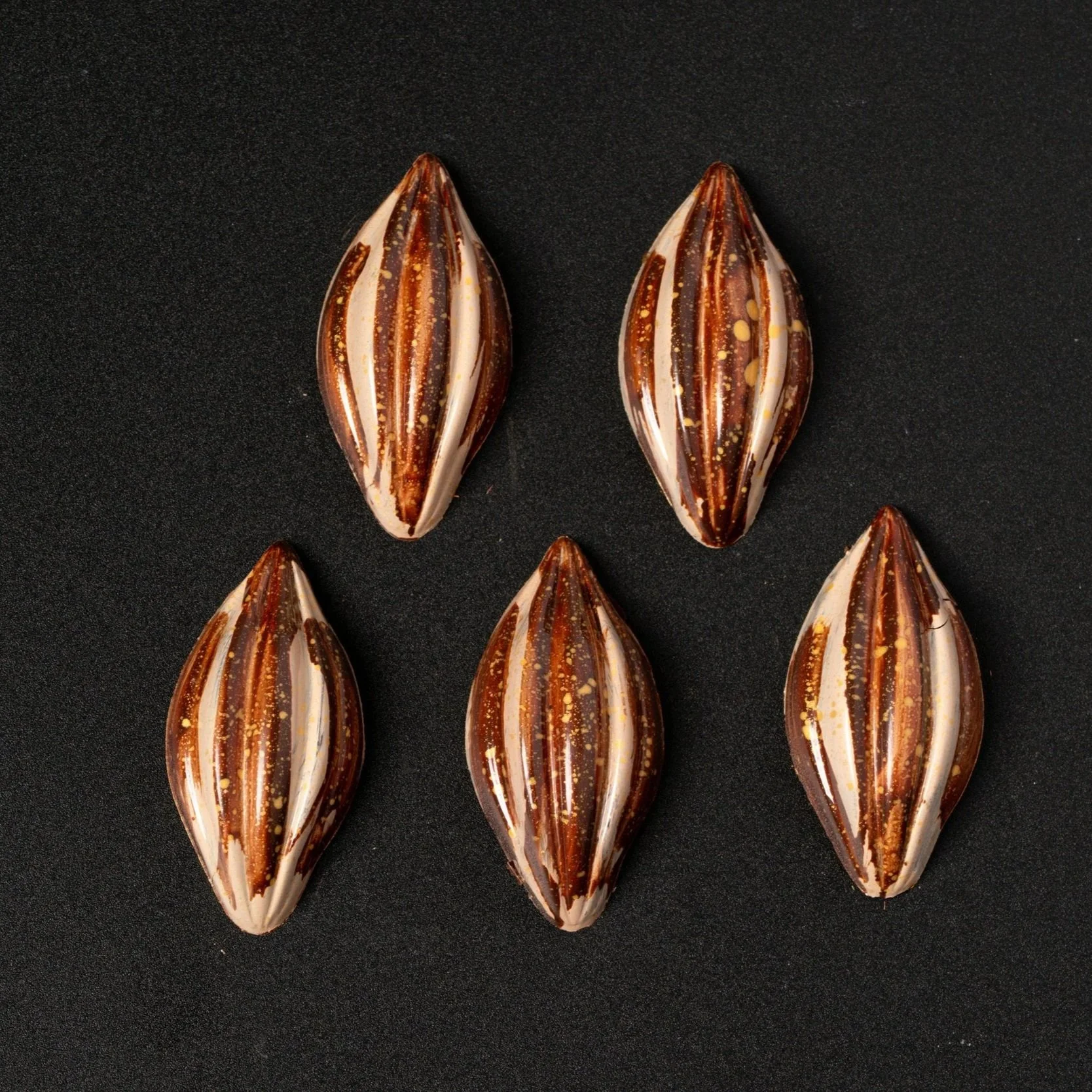 Five decorative teardrop-shaped chocolate bonbons with brown, cream, and gold swirls on a black background.